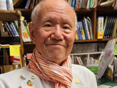 Manga artist Tetsuya Chiba smiling in his studio, wearing a white jacket and an orange-striped scarf in front of shelves of books and marker pens.