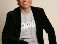 Portrait of Nintendo designer Shigeru Miyamoto smiling while seated, wearing a black velvet jacket over a grey T-shirt printed with Mii characters.